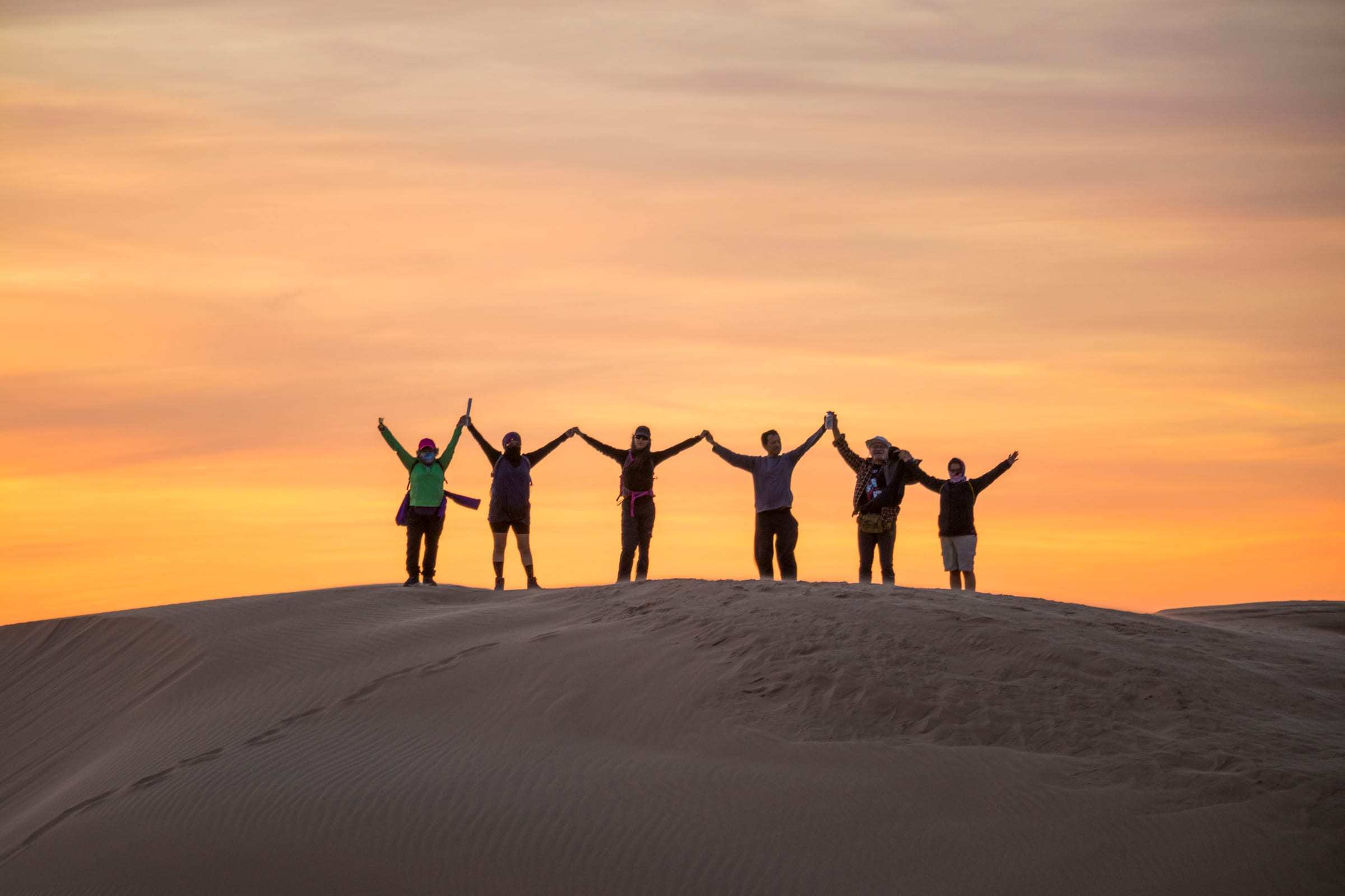Grupo de viajeros con los brazos arriba en la cresta de una duna al atardecer